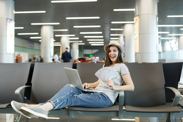 Молодая туристка-путешественница, работающая на ноутбуке, ждет в вестибюле международного аэропорта связку денег наличными. Пассажир, выезжающий за границу в выходные. Концепция полета
 - Фото, изображение