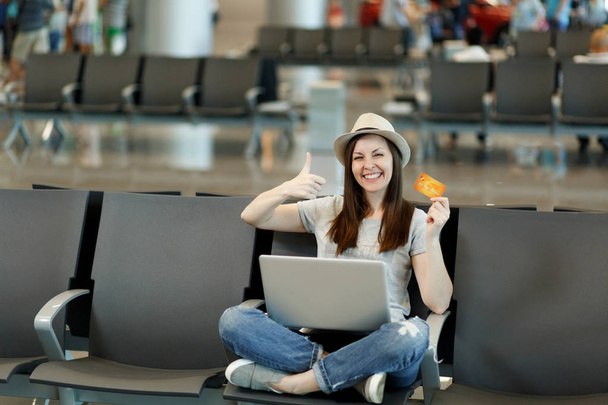 Viajante alegre turista mulher com laptop sentado com pernas cruzadas, mostrando polegar para cima segurar cartão de crédito esperar no hall de entrada no aeroporto. Passageiro viajando para o exterior em fuga de fim de semana. Conceito de voo aéreo
 - Foto, Imagem