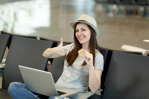Mladá usměvavá cestovatel turistické žena v klobouku sedět, pracovat na notebooku, držení bitcoin, ukazuje palcem, čekat v lobby hale na letišti. Cestující cestující do zahraničí na víkend. Koncept letu vzduchem - Fotografie, Obrázek