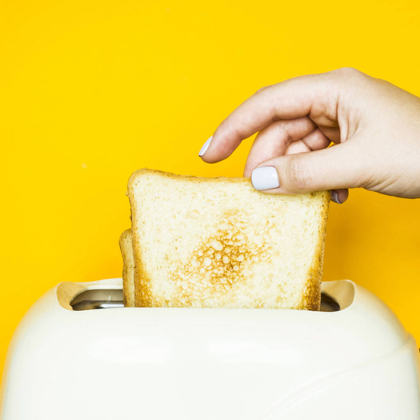 Toasted bread sticks out of the toaster on a yellow background. A woman's hand takes out a piece of bread - Zdjęcie, obraz