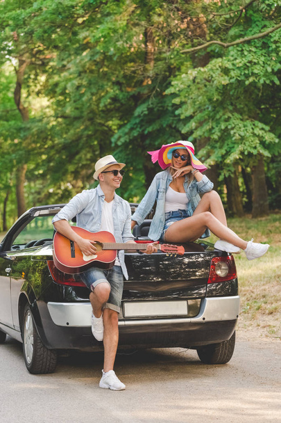Young fashionable couple playing acoustic guitar on the back side of their convertible car in nature. - Foto, immagini