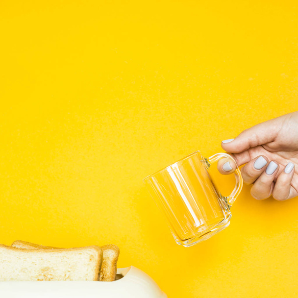 Toasted bread sticks out of the toaster on a yellow background. A woman's hand holds an empty cup for coffee. Breakfast concept - Φωτογραφία, εικόνα