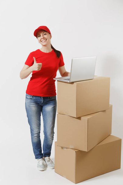 Ganzkörperporträt der Lieferantin mit roter Mütze, T-Shirt auf weißem Hintergrund. Kurierin steht neben leeren Kartons mit Laptop-Computer. Empfangspaket. Kopierraum - Foto, Bild