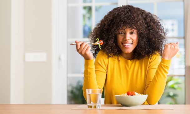 Mujer afroamericana comiendo ensalada de pasta en casa gritando orgullosa y celebrando la victoria y el éxito muy emocionada, animando emoción
 - Foto, imagen