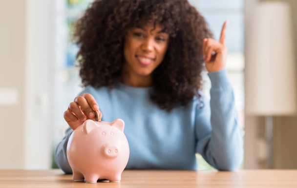 Mujer afroamericana ahorra dinero en alcancía sorprendida con una idea o pregunta apuntando con el dedo con la cara feliz, número uno
 - Foto, Imagen