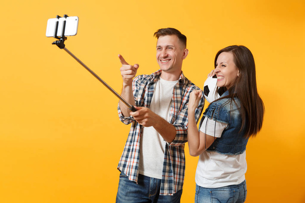Jeune homme de couple femme, fans de football faisant selfie sur téléphone mobile avec bâton égoïste monopode, remonter le moral de l'équipe de soutien, ballon de football isolé sur fond jaune. Sport famille loisirs style de vie concept
 - Photo, image