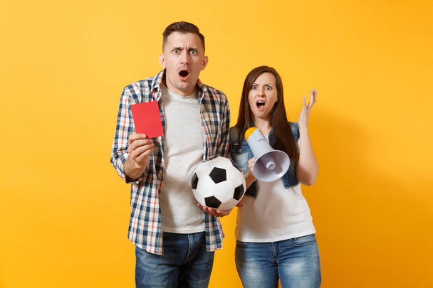 Angry fun expessive crazy couple, woman man football fans screaming, cheer up support team with soccer ball, megaphone, red card isolated on yellow background. Sport family leisure lifestyle concept - 写真・画像