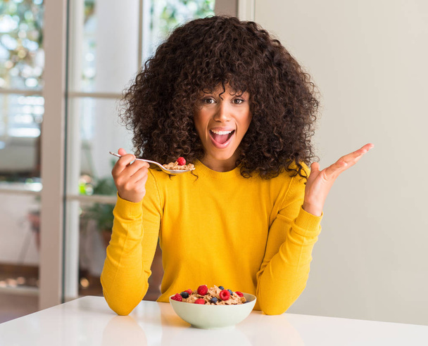 African american woman eating cereals, raspberries and blueberries very happy and excited, winner expression celebrating victory screaming with big smile and raised hands - Photo, Image