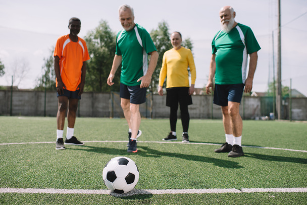 selective focus of football ball and multicultural elderly men on green football field - Photo, Image