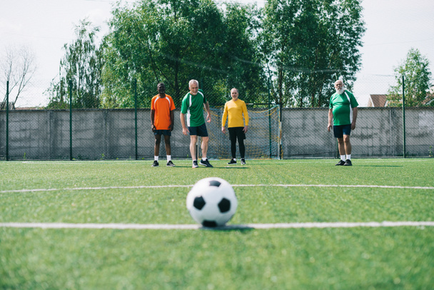 selective focus of football ball and multicultural elderly men on green football field - Foto, imagen