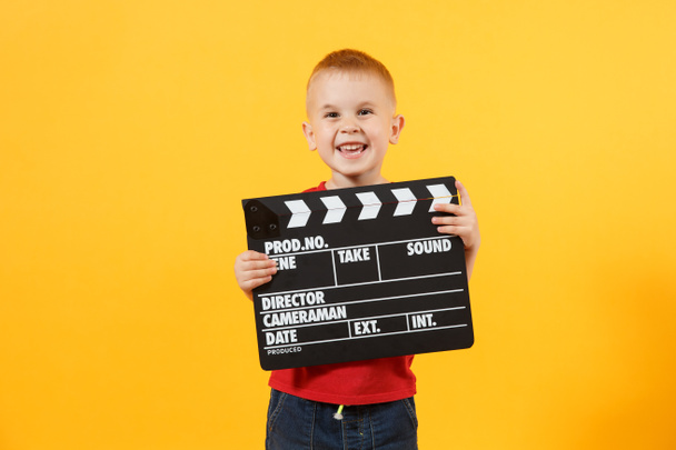 Little cute kid baby boy 3-4 years old in red t-shirt holding in hand classik black film making clapperboard looking camera isolated on yellow background. Kids childhood lifestyle concept. Copy space - Foto, Bild