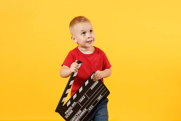 Little cute kid baby boy 3-4 years old in red t-shirt holding in hand classik black film making clapperboard looking camera isolated on yellow background. Kids childhood lifestyle concept. Copy space - Valokuva, kuva