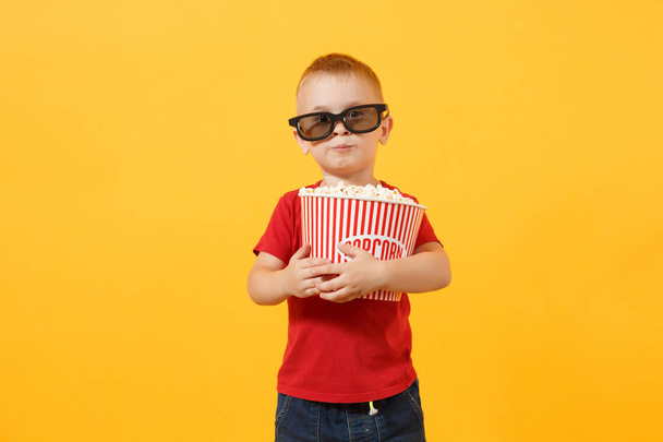 Little cute kid baby boy 3-4 years old in red t-shirt, 3d imax cinema glasses holding bucket for popcorn, eating fast food isolated on yellow background. Kids childhood lifestyle concept. Copy space - Foto, Imagen