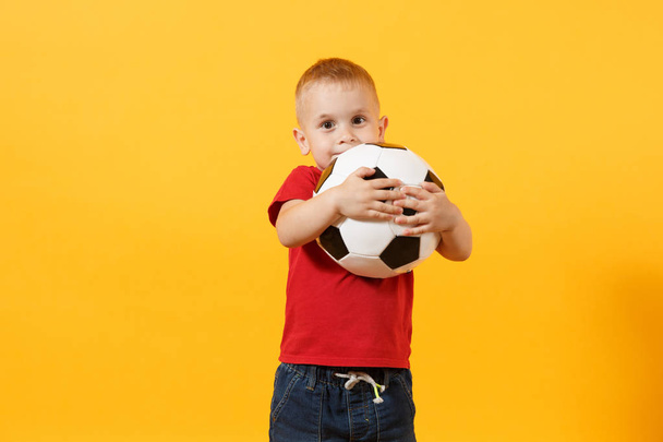Little cute kid baby boy 3-4 years old, football fan in red t-shirt holding in hand soccer ball isolated on yellow background. Kids sport family leisure lifestyle concept. Copy space advertisement - Фото, изображение