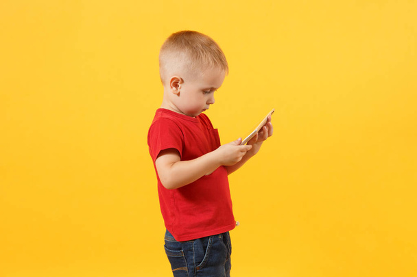 Little cute kid baby boy 3-4 years old in red t-shirt holding in hand tablet pc computer isolated on yellow background. Kids childhood lifestyle concept. Problem of children and gadgets. Copy space - Photo, image