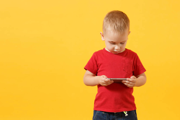 Little baby boy 3-4 years old in red t-shirt holding in hand, playing game on mobile phone isolated on yellow background. Kids childhood lifestyle concept. Problem of children and gadgets. Copy space - Photo, Image