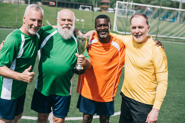 multiethnic smiling old sportsmen with champions cup standing on football field - Photo, Image