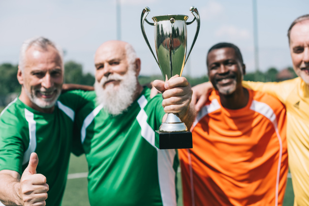 selective focus of multiethnic smiling old sportsmen with champions cup standing on football field - Photo, Image