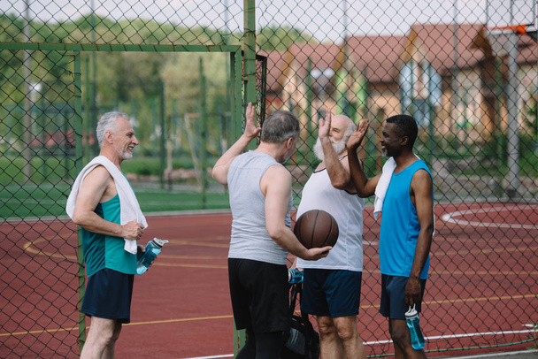 Ryhmä monirotuisia vanhoja urheilijoita, joilla on koripallo, antavat toisilleen leikkikentällä ylävitosen
 - Valokuva, kuva