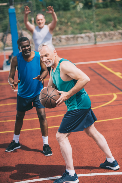valikoiva painopiste vanha mies koripallo käsissä ja monirotuinen ystäviä takana leikkikentällä
 - Valokuva, kuva