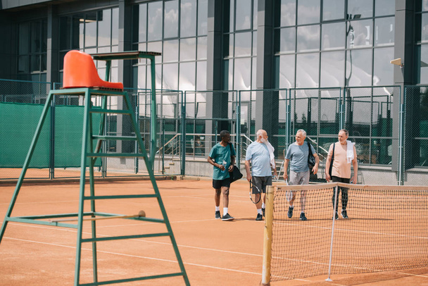 multiracial elderly friends with tennis equipment walking on court - Photo, Image