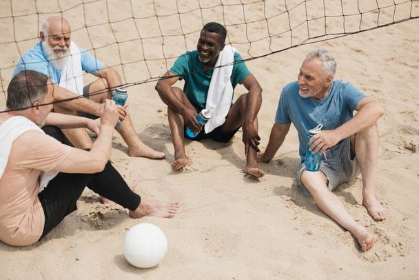 çok ırklı gülümseyen yaşlı voleybol oyuncular ile sportif su şişeleri maçtan sonra dinlenme - Fotoğraf, Görsel