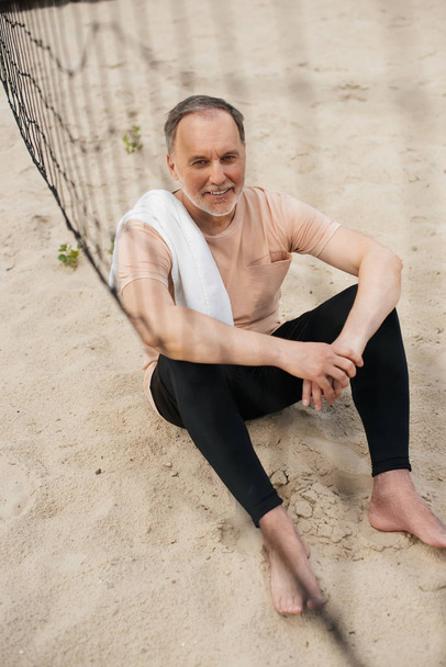 smiling elderly man with towel resting near net on sandy beach after playing volleyball - Zdjęcie, obraz