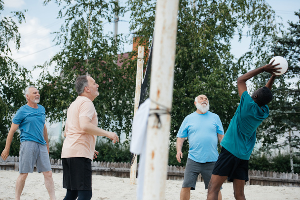 side view of multicultural old friends playing volleyball on beach on summer day - Foto, Imagen