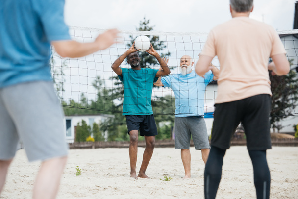 vista parziale di vecchi amici multiculturali che giocano a pallavolo sulla spiaggia durante la giornata estiva
 - Foto, immagini