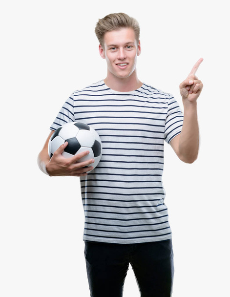 Joven guapo rubio sosteniendo pelota de fútbol muy feliz apuntando con la mano y el dedo hacia un lado
 - Foto, Imagen