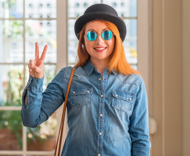 Elegante donna rossa con cappello a bombetta e occhiali da sole che mostra e indica con le dita il numero due sorridendo fiducioso e felice
. - Foto, immagini