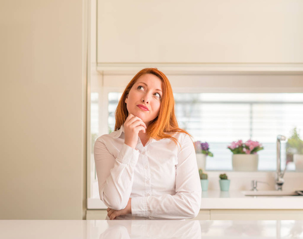 Redhead woman at kitchen with hand on chin thinking about question, pensive expression. Smiling with thoughtful face. Doubt concept. - Photo, Image