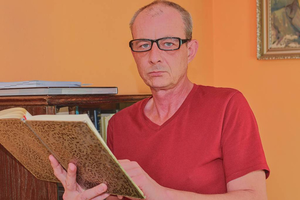 Portrait of middle aged man. Man is reading book in living room. Mature man is standing next to bookcase.  - Photo, Image