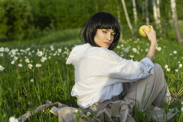 Beautiful stylish girl wearing a trendy white shirt, beige trousers and a black wig holds a green apple in sunlight sitting on a green meadow grass. Advertising and fashion design. Copy space. - Photo, image