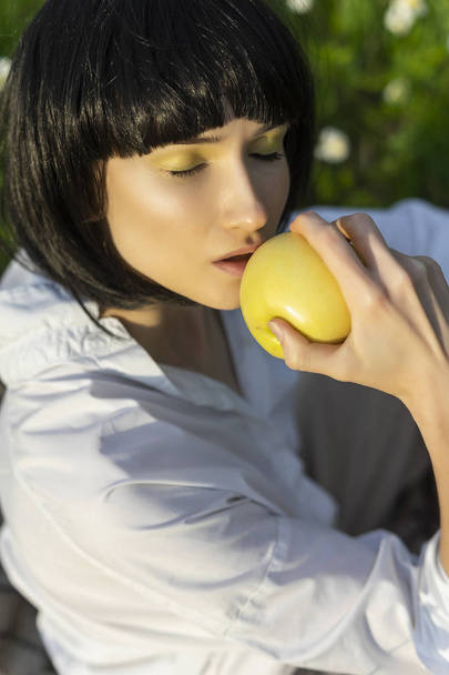 Beautiful stylish girl wearing a trendy white shirt, beige trousers and a black wig holds a green apple in sunlight sitting on a green meadow grass. Advertising and fashion design. Copy space. - Фото, зображення