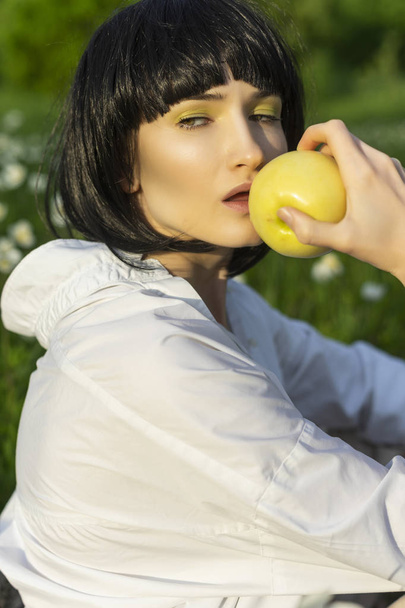 Beautiful stylish girl wearing a trendy white shirt, beige trousers and a black wig holds a green apple in sunlight sitting on a green meadow grass. Advertising and fashion design. Copy space. - Photo, Image