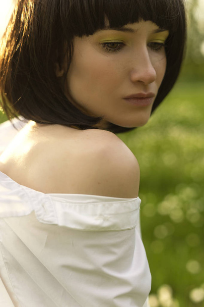 Art style close up photo of a girl with naked shoulder wearing a trendy white shirt and a black wig in sunset on a background of green meadow. Advertising and fashion design. Copy space - Foto, Bild