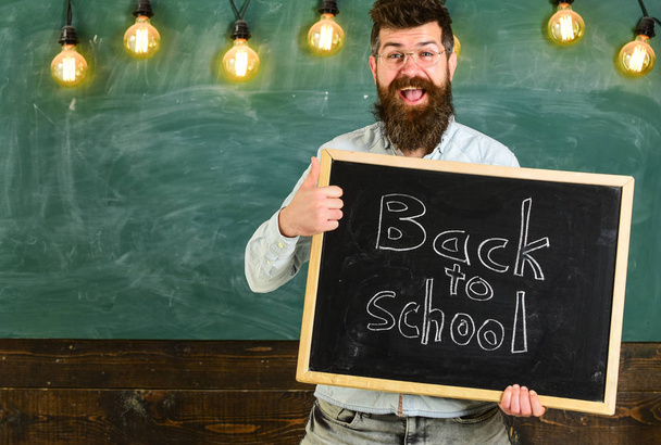 Teacher in eyeglasses holds blackboard with inscription back to school. Man with beard on happy face welcomes students with thumb up gesture, chalkboard on background. Back to school concept - Фото, изображение