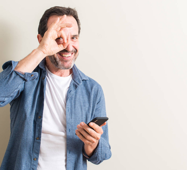 Uomo anziano utilizzando smartphone con volto felice sorridente facendo segno ok con mano sugli occhi guardando attraverso le dita
 - Foto, immagini