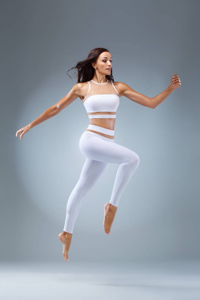 Attractive excited fitness girl, ballerina dancer in sportwear jumping of joy isolated over gray background. - Фото, изображение
