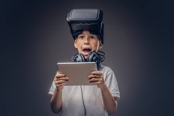 Portret van een verbaasd kleine jongen gekleed in een wit t-shirt, met VR-bril en koptelefoon houdt een digitaal tablet in een studio, geïsoleerd op donkere achtergrond. Onderwijs en - Foto, afbeelding