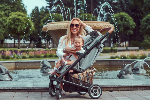 Beyaz bir bluz, şort ve bir bebek arabası bir çeşme şehir Parkı yakınındaki küçük kızıyla birlikte poz güneş gözlüğü mutlu sarışın kadın giyinmiş. - Fotoğraf, Görsel