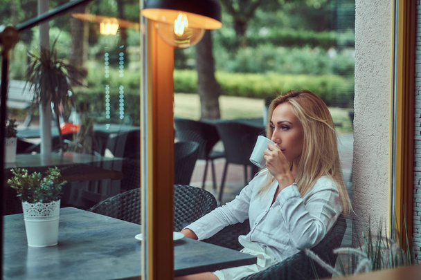 Portrait of a beautiful blonde female dressed in a white blouse sitting at a table drinks coffee at terrace cafe. - Foto, Imagem