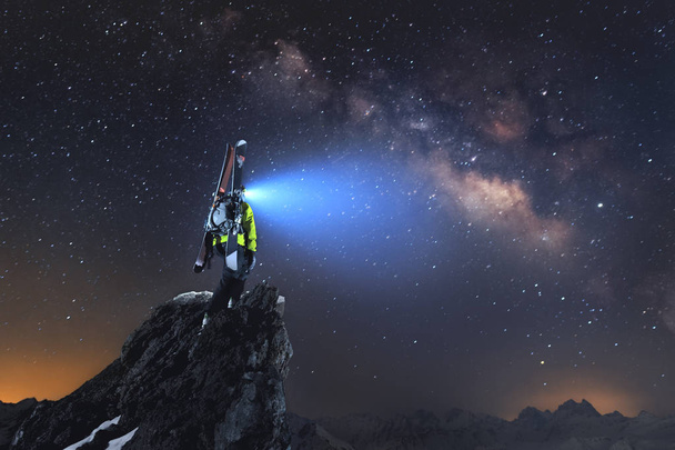 Ночной пейзаж. Профессиональный лыжник с рюкзаком и лыжами стоит на скале в горах и светит фарой в Млечном Пути
 - Фото, изображение