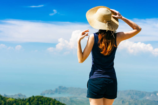 Young traveling woman wearing hat and standing on the top of the mountain cliff watching beautiful view of woods and blue sky and clouds on vacation. - Foto, imagen