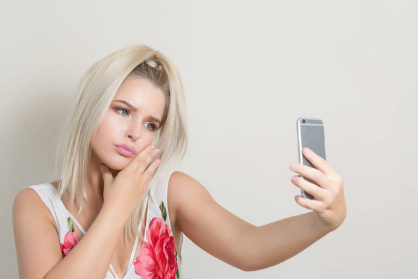 灰色の背景に対して携帯電話上で selfie を稼ぐ感情的な金髪女性 - 写真・画像