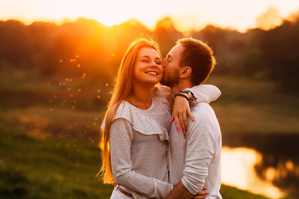 A guy with a beard gently kisses his girlfriend on a cheek and she is enjoying it. gentle hugs on the background of the sunset - Photo, Image
