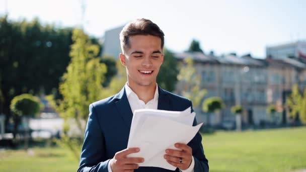 Young happy businessman in a stylish suit carefully examines the papers outdoor. Successful lifestyle, contract, responsibility. Business, people, paperwork conception - Filmmaterial, Video