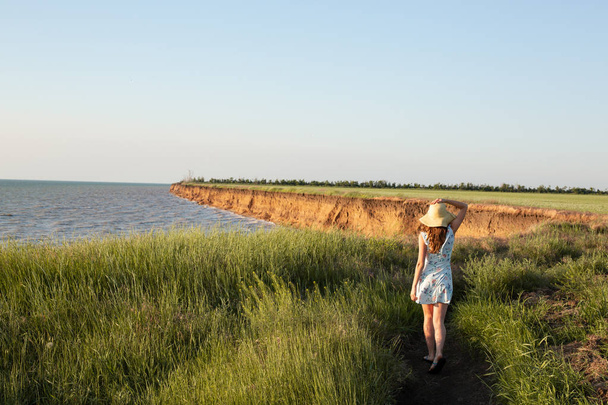 Young Romantic Woman in Blue Dress and Hat Walking the Beautiful Field against The Sea at Sunset. Standing Back to Camera. A Girl with Red  Hair. Summer Time - Foto, Bild