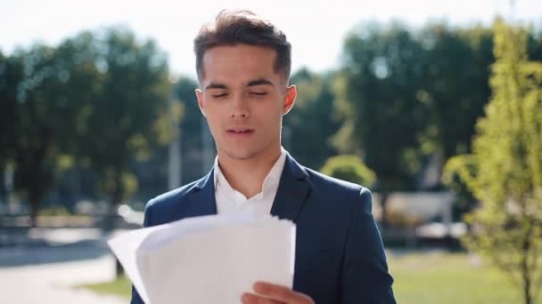 Young happy businessman in a stylish suit carefully examines the papers outdoor. Successful lifestyle, contract, responsibility. Business, people, paperwork conception - Metraje, vídeo